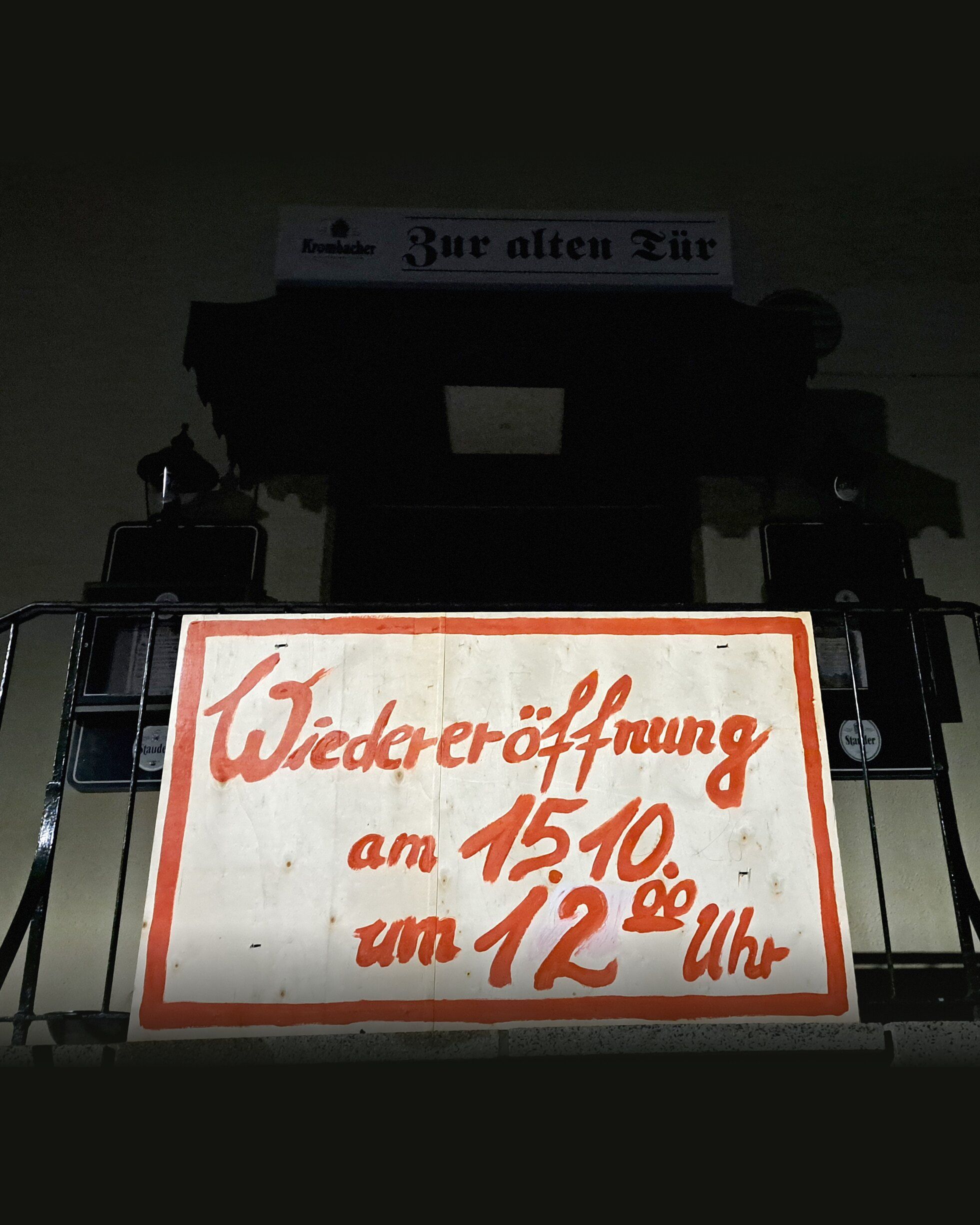Eingang bei Nacht: Leuchtschild Zur alten Tür über der Treppe, davor ein gemaltes Plakat: Wiedereröffnung am 15.10. um 12 Uhr
